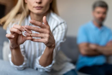 Divorce amiable : procédure, délais et coûts du divorce sans juge Femme enlevant son alliance avec son mari en arrière plan flouté, bras croisés, illustrant un divorce amiable