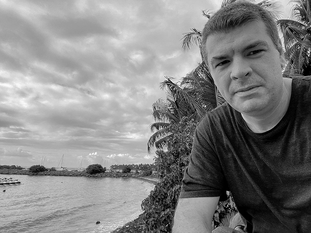 Nicolas, photo de l'auteur en noir et blanc en t-shirt au bord d'une baie de Guadeloupe, avec des cocotiers, des voiliers au mouillage et un ciel chargé de nuages